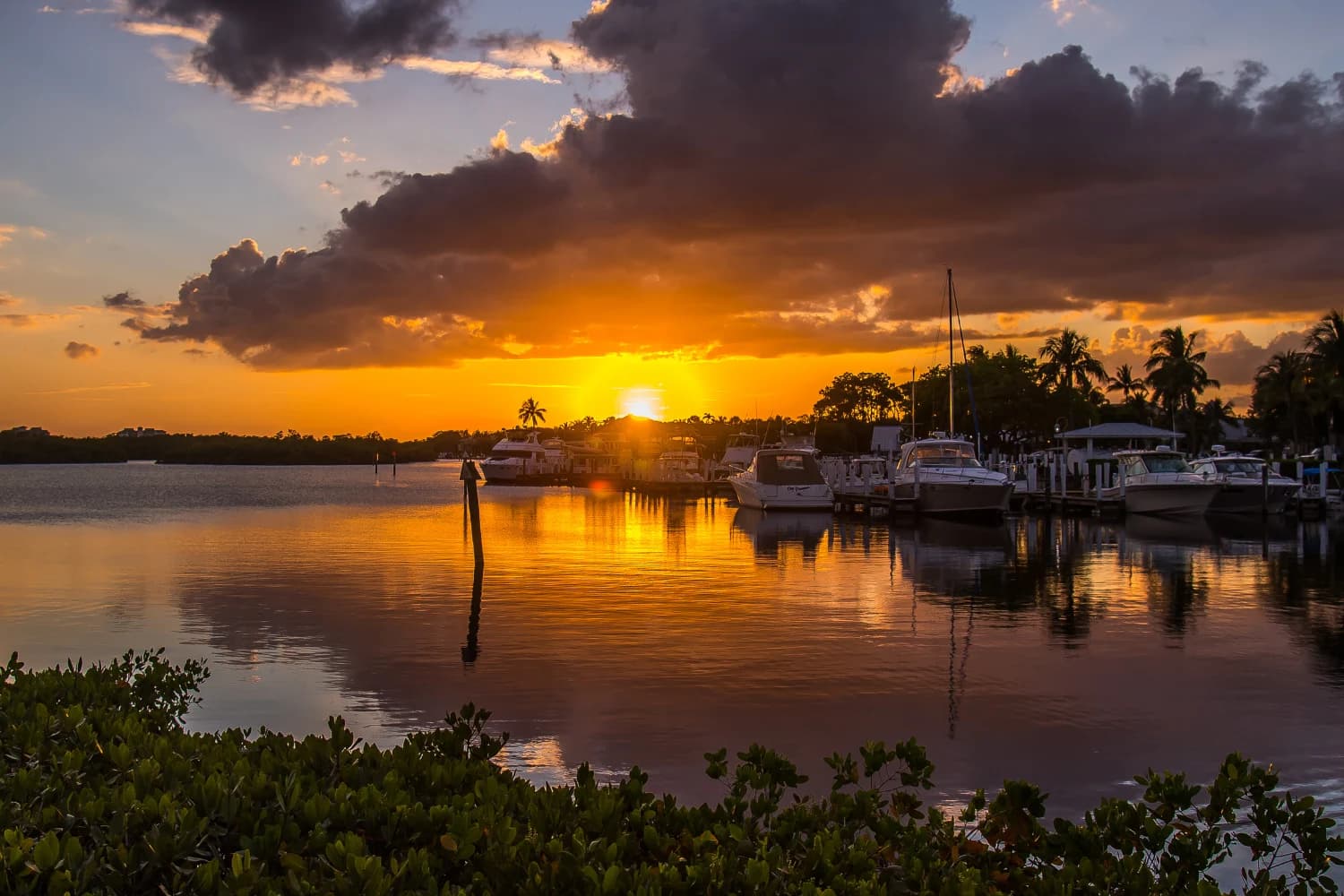 Sunset over a calm marina with docked boats and reflections on the water, framed by dark clouds and shoreline greenery.