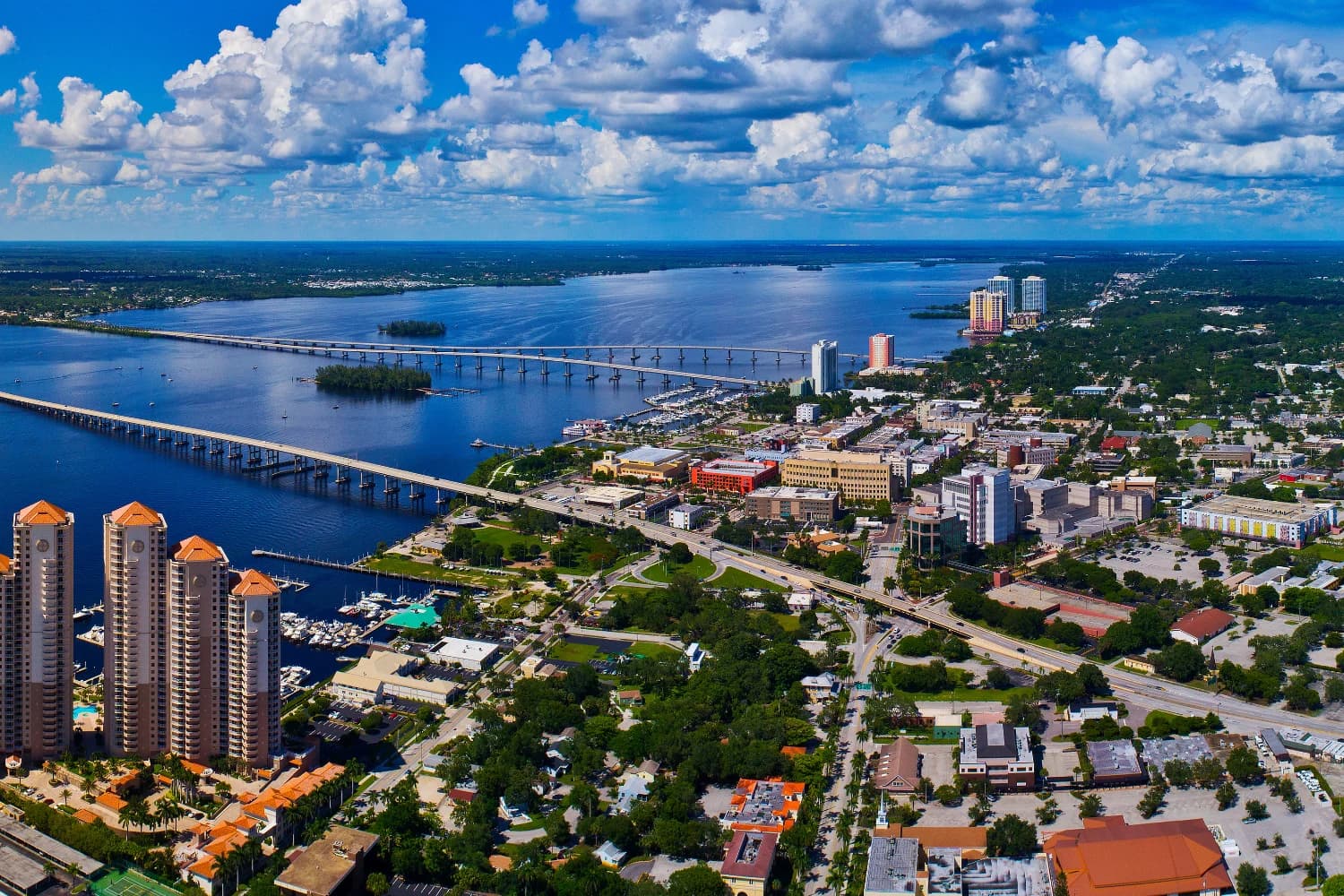Aerial view of a coastal city with long bridges spanning wide water, marinas and high-rise buildings along the waterfront under scattered clouds.