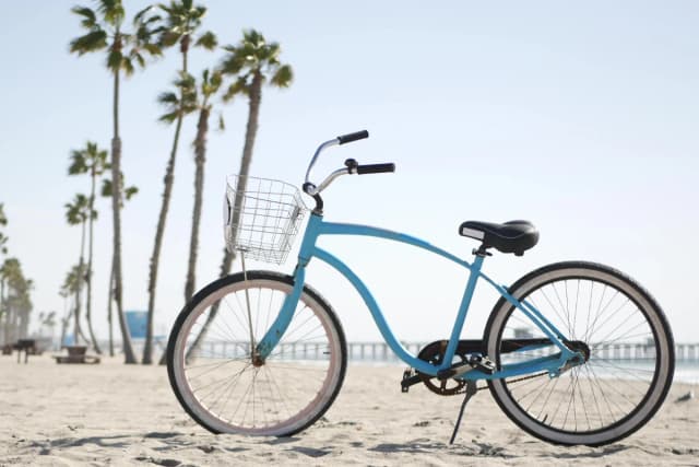 Blue beach cruiser bicycle with a front basket parked on sand with tall palm trees in the background.