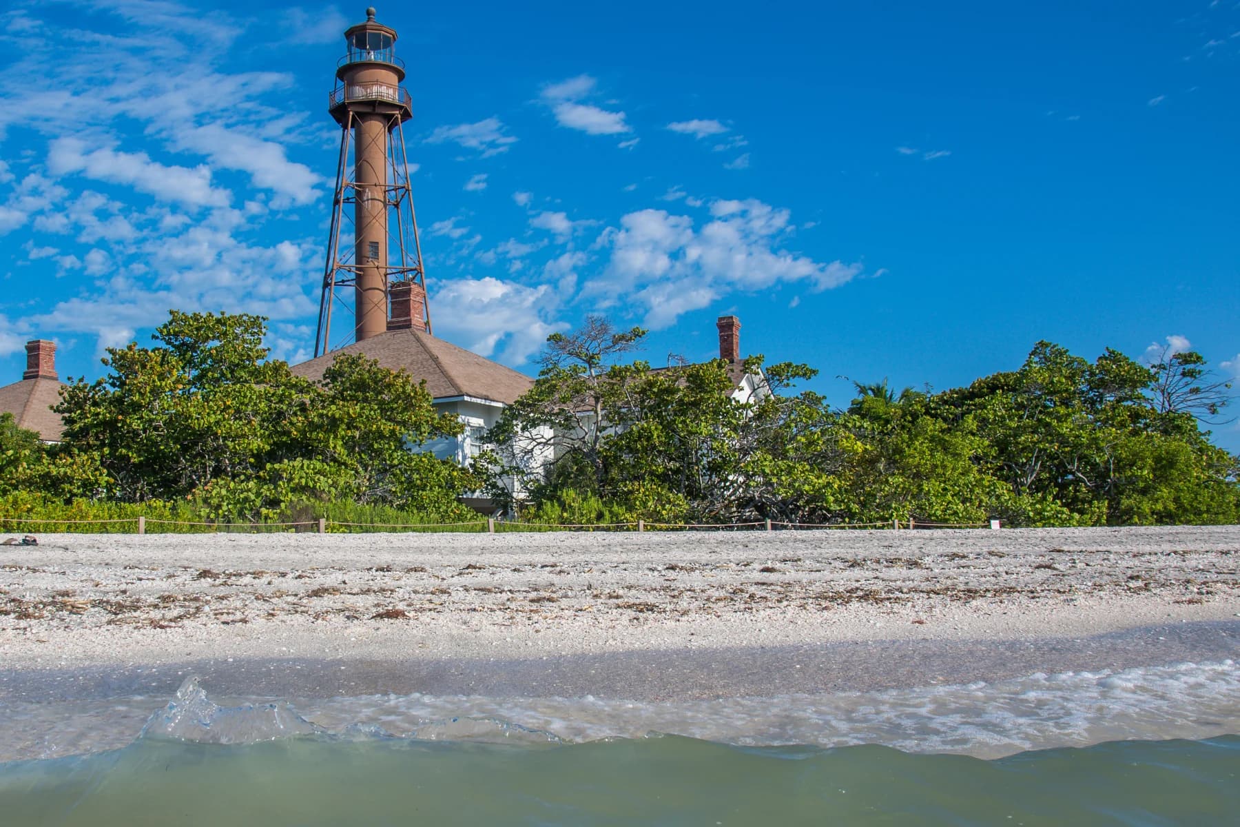 Tall lighthouse rising above coastal greenery, viewed from the shoreline with shallow water in the foreground under a bright blue sky.