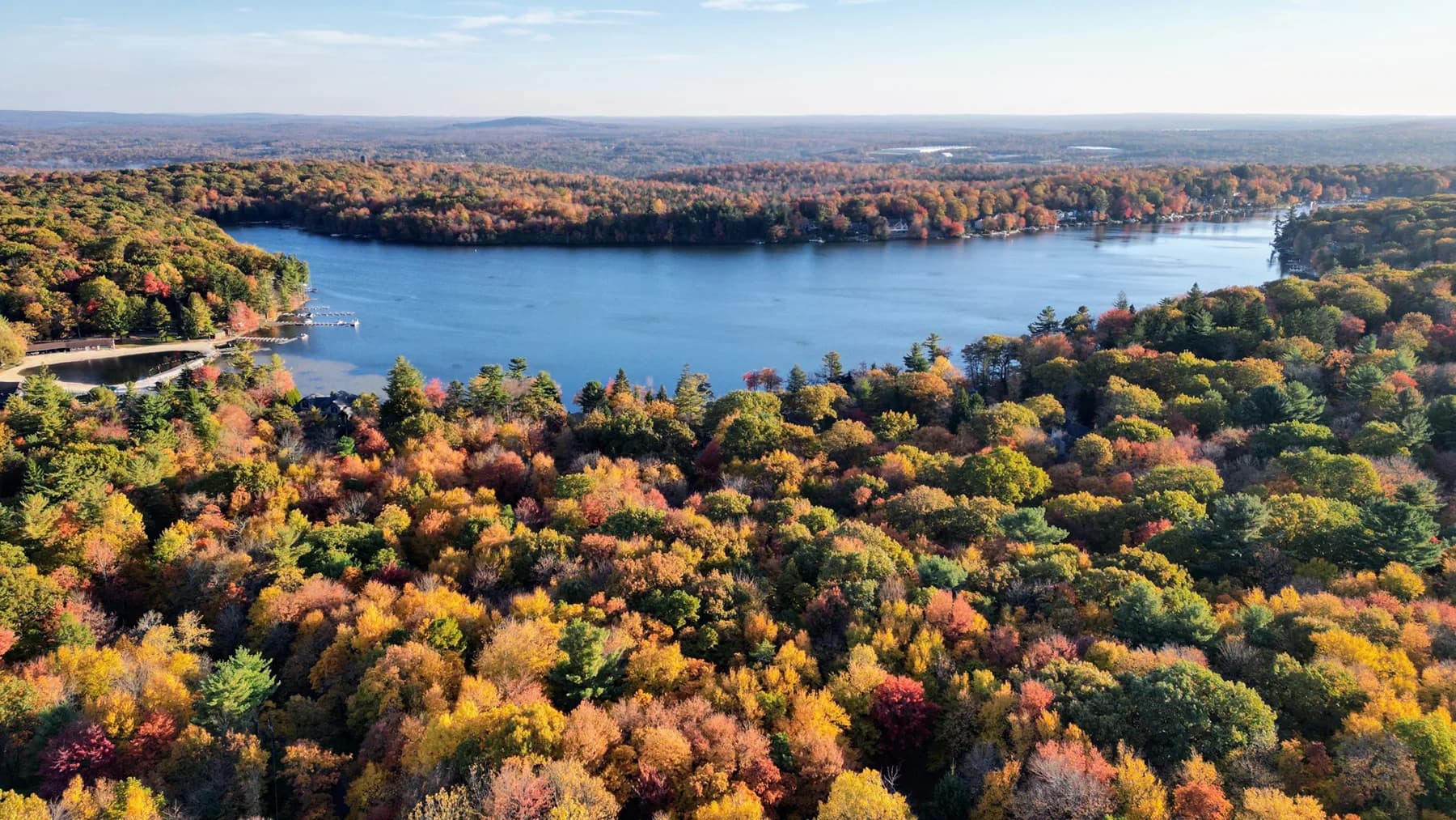 “Aerial view of a large lake surrounded by dense autumn forest, with vibrant fall colors of red, orange, yellow, and green stretching across rolling hills under a bright, clear sky