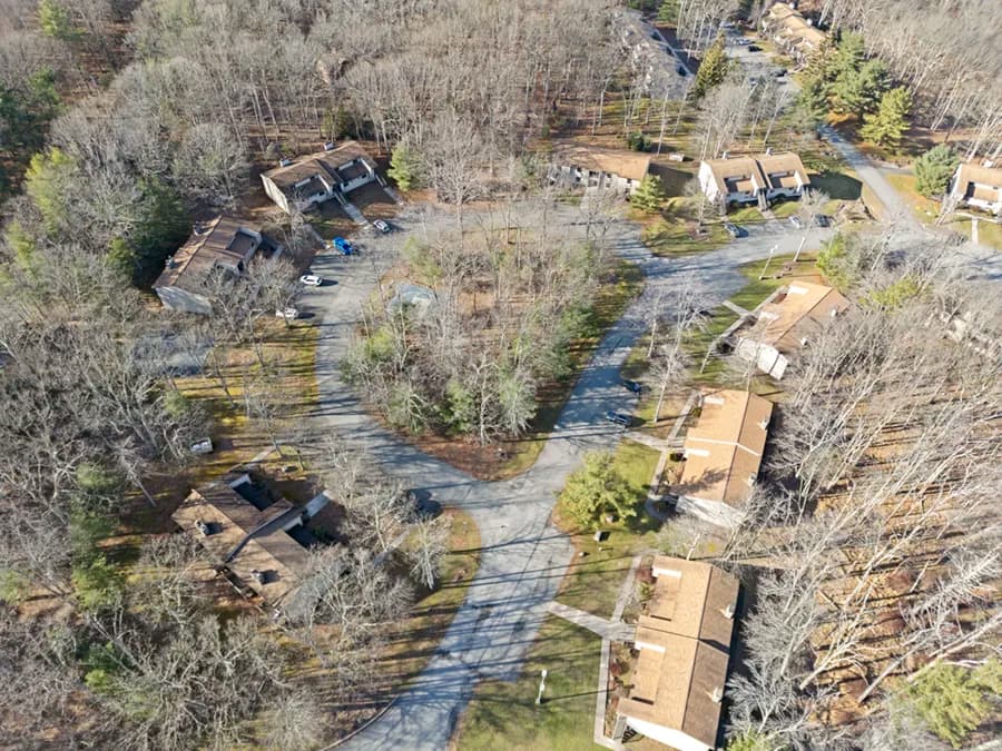 High-angle aerial view of a wooded residential community with multiple tan-roofed townhome buildings arranged along curving roads, surrounded by bare winter trees and scattered evergreens.