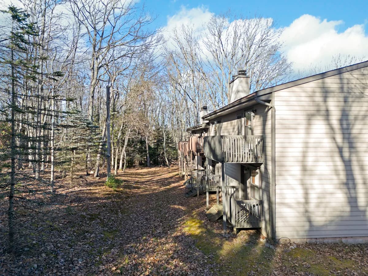 Side view of a beige wood-sided residential building with balconies overlooking a forested area filled with bare trees and evergreen pines under a bright, partly cloudy sky.