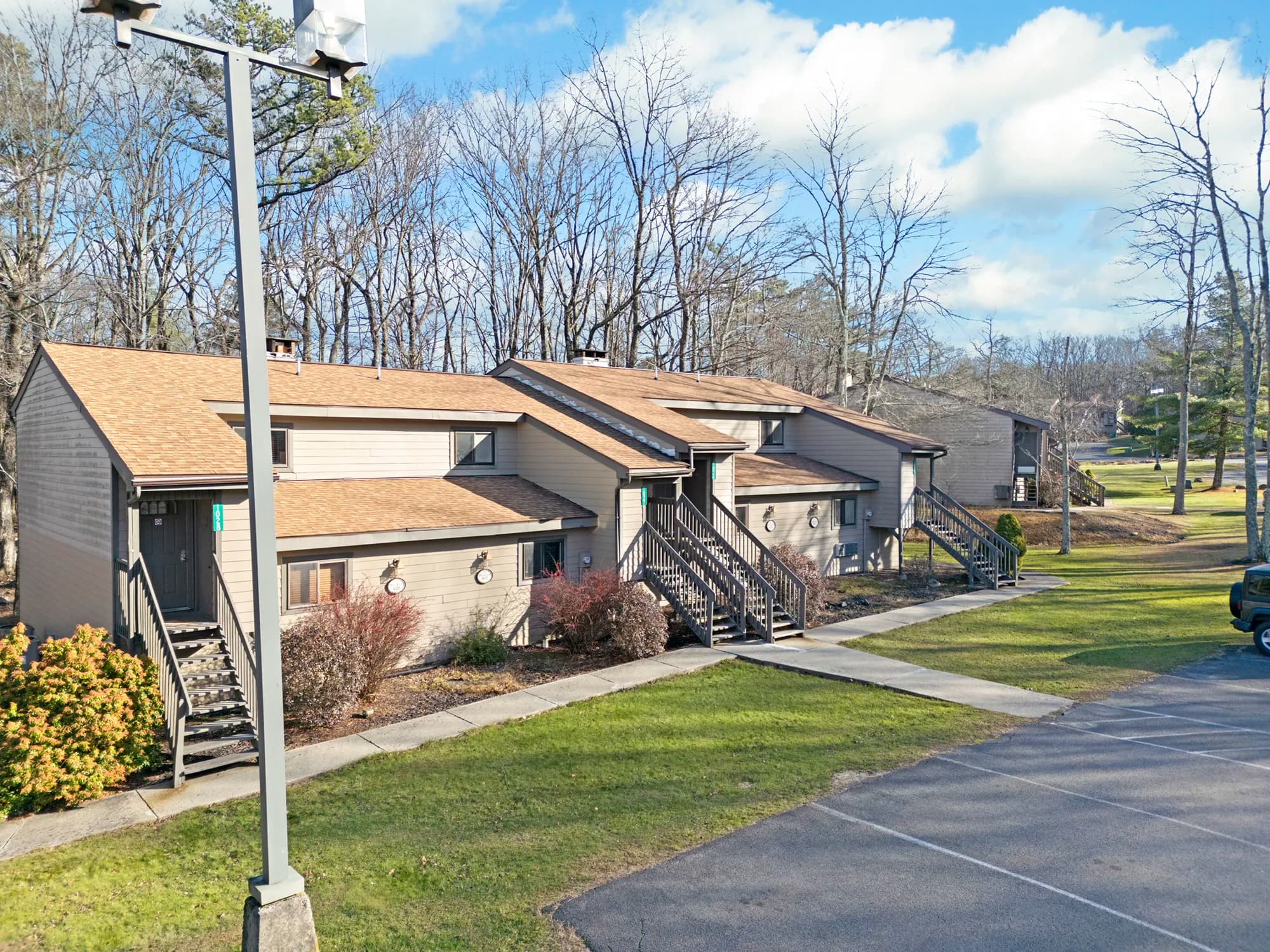 Row of modern tan wood-sided townhome units with exterior staircases, surrounded by early-spring trees and open grassy areas, with a parking lot and lamppost in the foreground under a bright partly cloudy sky.